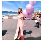 A woman in a blush satin luxury fashion tube dress with a high slit holds light pink balloons on a boardwalk