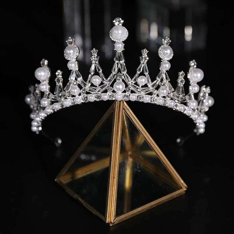 A sparkling silver bridal crown rhinestone tiara with clear crystals and white pearls displayed on a gold stand