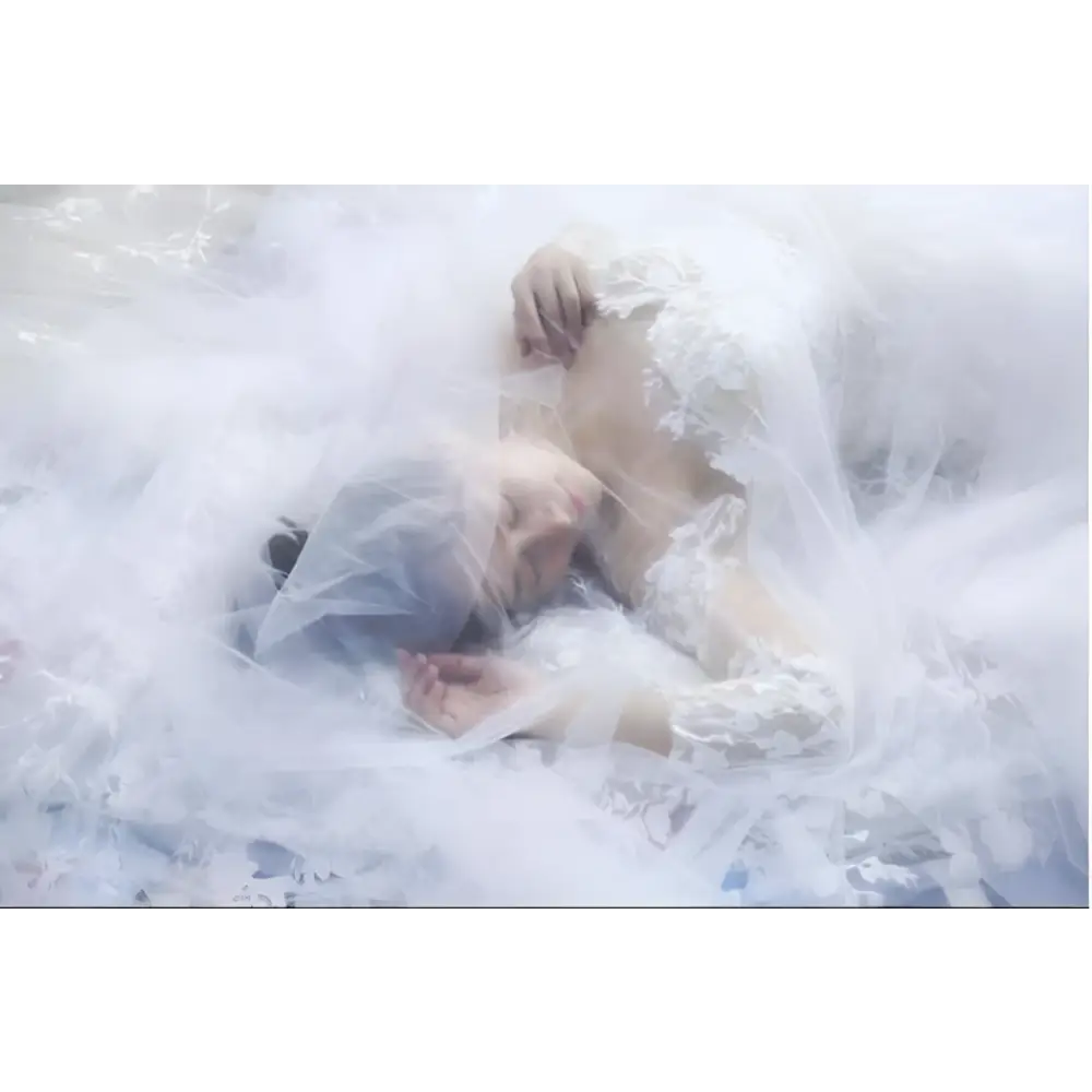 A woman lies entwined in a sheer white lace bridal wedding veil with floral embroidery and sheer tulle fabric