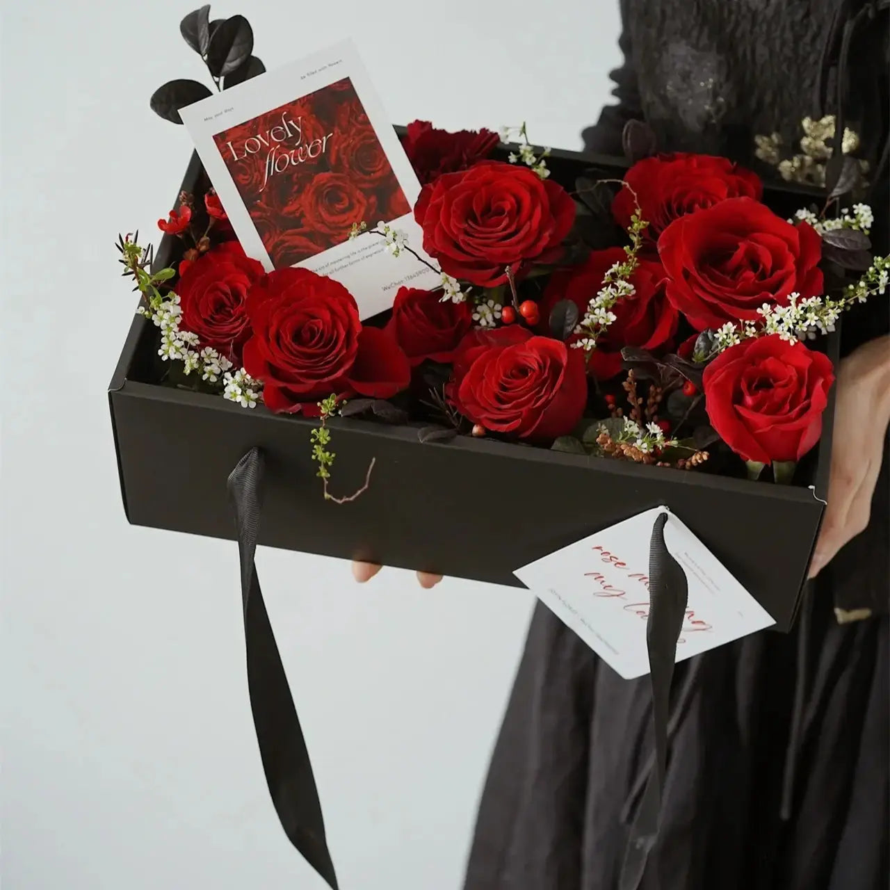 Elegant black rectangular handheld flower box with a clear window filled with vibrant red roses and delicate white filler flowers featuring a red bow knot handheld design.
