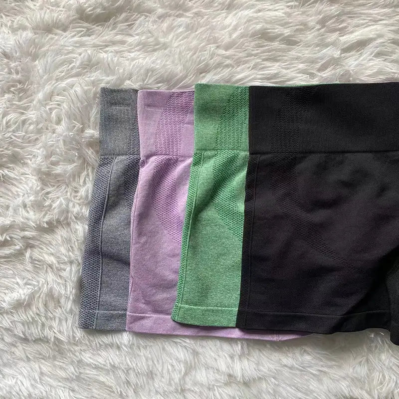 Four pairs of women sport seamless high-waisted leggings in gray, lavender, green, and black are neatly displayed on a fluffy white surface as sport seamless short leggings.