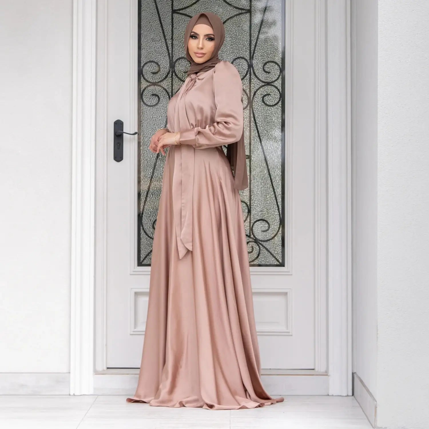 a flowing dusty rose-colored large hem satin dress with long sleeves and a tied waistband designed as a long sleeve abaya in fashion large hem style
