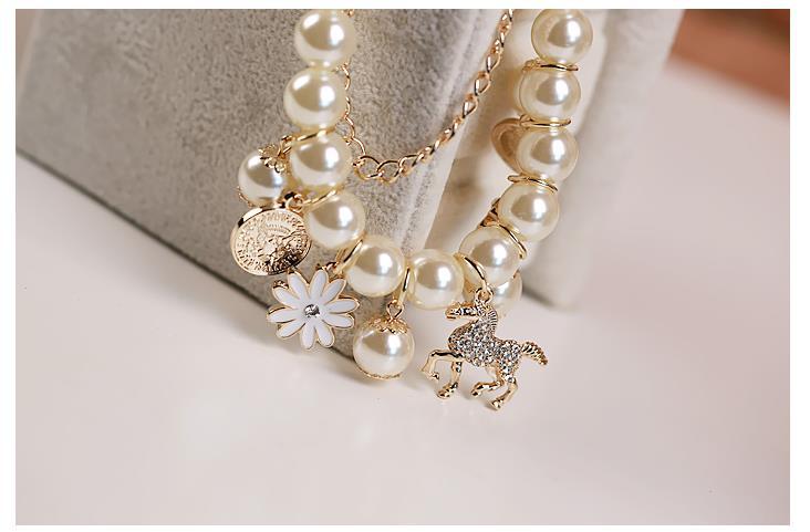 A delicate gold-toned pearl flower bracelet featuring cream-colored pearls and dangling charms including a white enamel daisy, a glittering gold horse figurine, and an ornate coin pendant.
