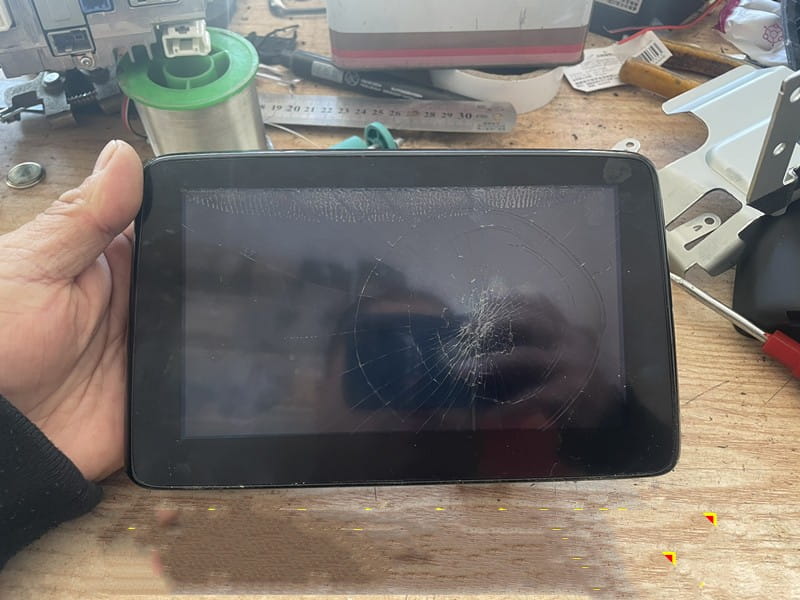 Black-framed original car exterior touch screen tablet with a cracked car exterior screen held in hand against a cluttered workbench background.