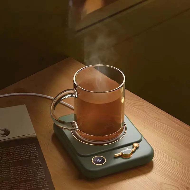 A sleek dark green milk heating insulation base desktop with a gold-toned temperature display and control knob holds a clear glass mug filled with steaming tea.
