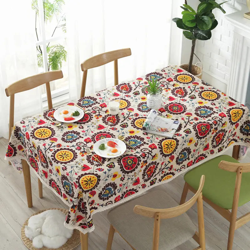 a vibrant linen table cloth featuring a colorful sunflower cotton floral and circular pattern in reds yellows blues and greens on an off-white background with lace trim