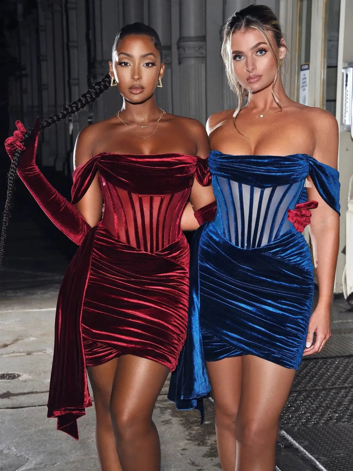 Two women wear off the shoulder velvet sexy evening dresses in deep red and royal blue highlighting a breast sexy evening style.