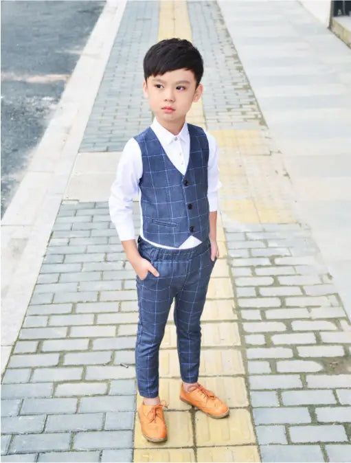 A young boy wearing a navy blue plaid children’s three-piece fashion suit with a comfort suit vest, white collared shirt, matching navy trousers, and tan leather shoes.