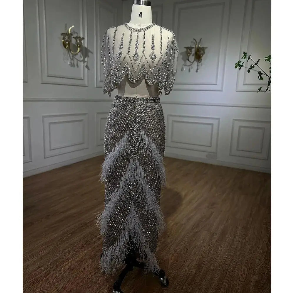 A shimmering silver nude two-piece evening dress with silver beaded details and feather accents on the skirt displayed on a mannequin.
