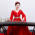 A woman in a vibrant red long guzheng performance dress with silver embroidery sits beside a dark wooden guqin