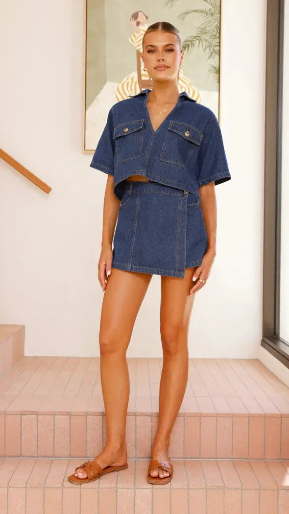 A woman wears a navy blue irregular denim short pantskirt suit featuring a short sleeve loose crop top with two front chest pockets and matching denim shorts.