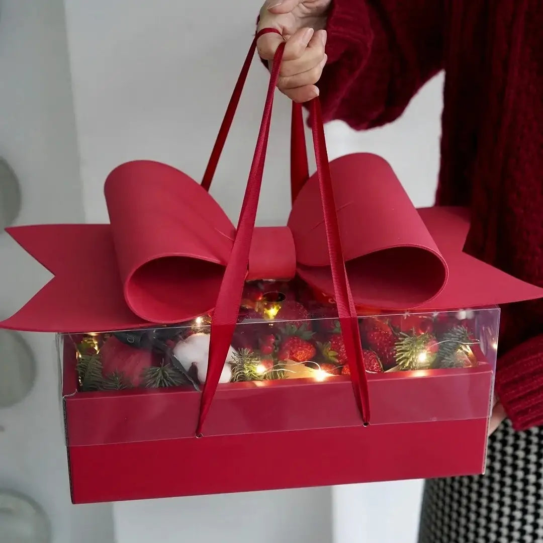 Red bow knot handheld flower box in red with a large glossy bow on top and clear sides revealing festive holiday decorations inside.