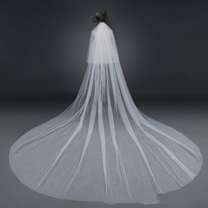 A flowing white bridal wedding veil widened tail made of delicate sheer tulle fabric featuring a small floral arrangement at the top.