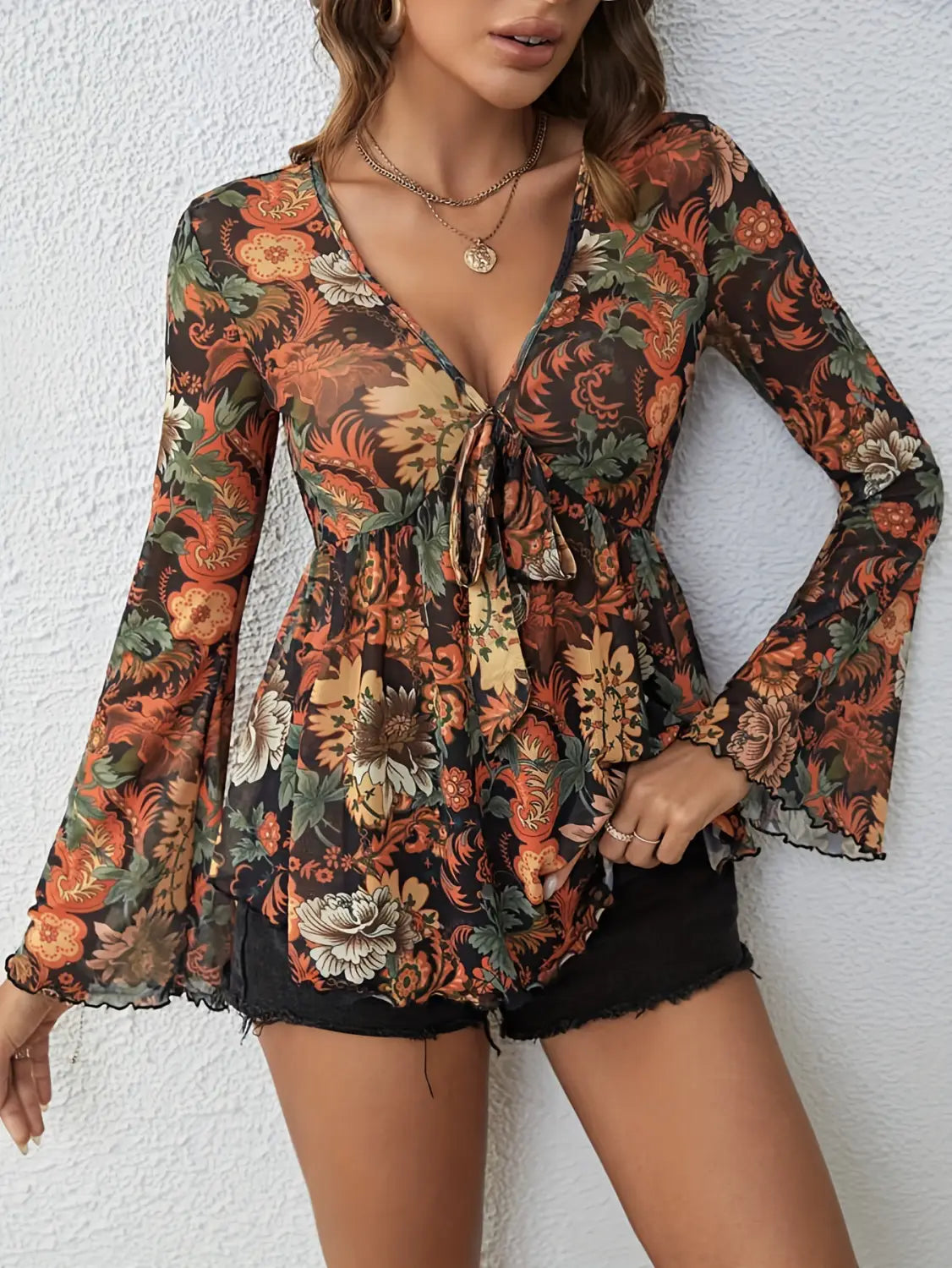A mesh printed knotted flare sleeve sheer long-sleeved top with a deep v-neck and tie-front detail features an intricate floral pattern in warm tones of orange rust cream and green on a dark brown background.