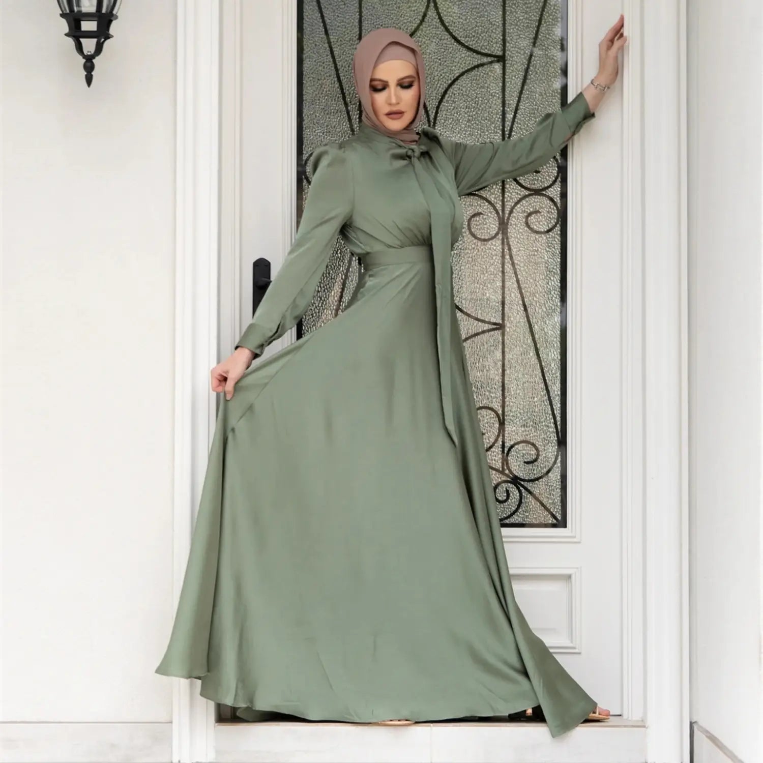 A flowing sage green satin maxi dress with large hem and long sleeves featuring a high neckline and an elegant bow at the waist in a large hem satin dress style.