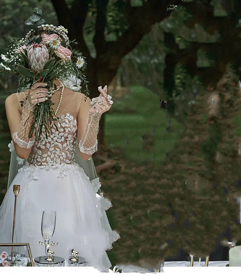 A bride in an elegant white lace wedding dress with a sheer veil and beaded embellishments holds a large bouquet obscuring her face in a lace summer couple photo shoot.