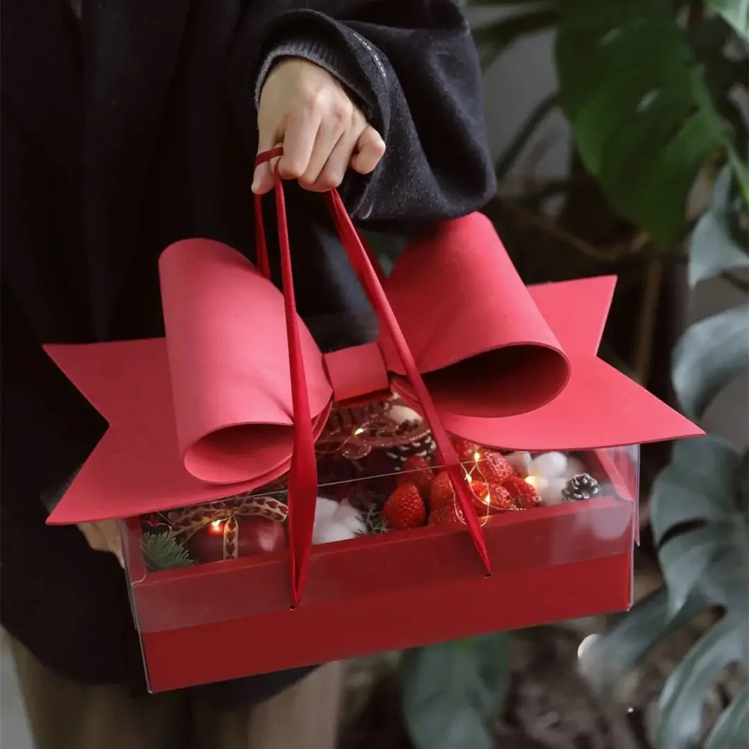A vibrant red bow knot handheld flower box with a large sculpted paper bow handle and clear window sides revealing festive contents.