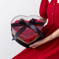 Heart shaped transparent flower packing box with vibrant red roses and a glossy black satin ribbon tie