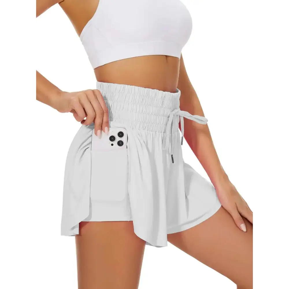 White high waist flowy athletic shorts with elastic waistband drawstring and side pockets holding a smartphone
