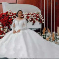 A woman in a white lace ball gown wedding dress with long sleeves and off the shoulder neckline sits gracefully