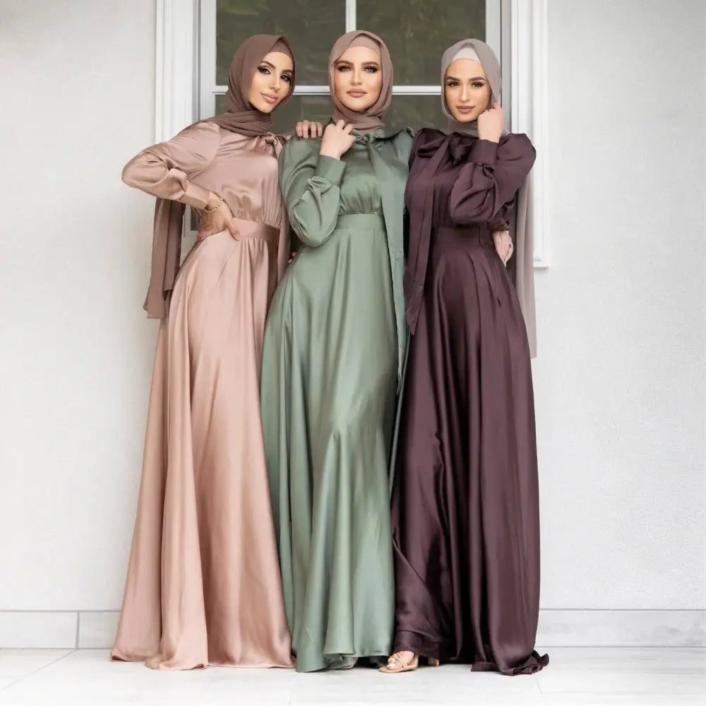 Three women wearing large hem satin long sleeve abayas in dusty rose sage green and deep burgundy colors