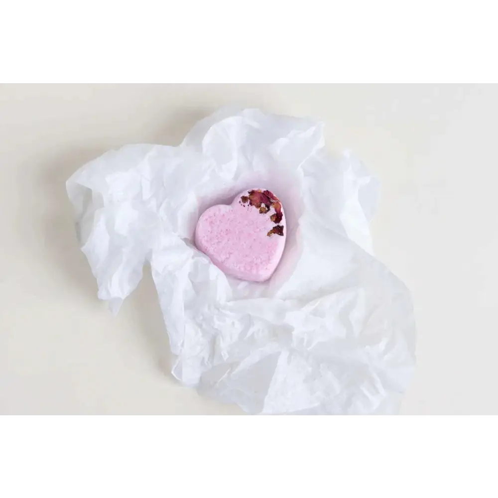 A soft pink heart-shaped bath bomb with dried rose petals in crinkled white tissue from lavender heart shower steamers gift set