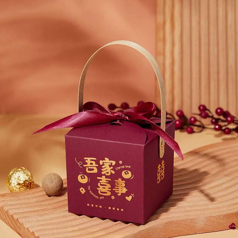 A maroon minimalist wedding candy gift box with gold foil lettering, a matching ribbon bow, and a light-colored handle