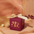 A maroon minimalist wedding candy gift box with gold foil lettering satin ribbon bow and gold handle for gifting