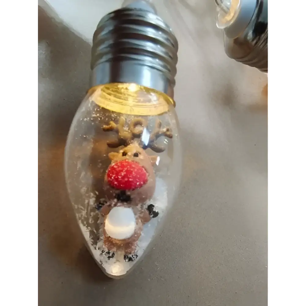 A clear glass christmas led string bulb ornament featuring a miniature scene reindeer with red nose and faux snow