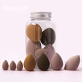 A set of earth-toned teardrop-shaped hydrophilic polyurethane beauty egg makeup sponges arranged around a clear jar
