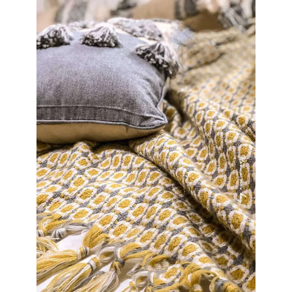 Gray cushion with textured fabric and black tassels paired with mustard yellow geometric knitted sofa blanket cover