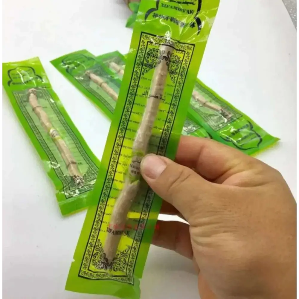 A hand holds a green-packaged natural miswak tooth cleaning stick with ornate black and gold arabic labels
