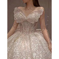 A shimmering off-white one-shoulder wedding dress with intricate beading, sequins, and a dramatic neckline bow