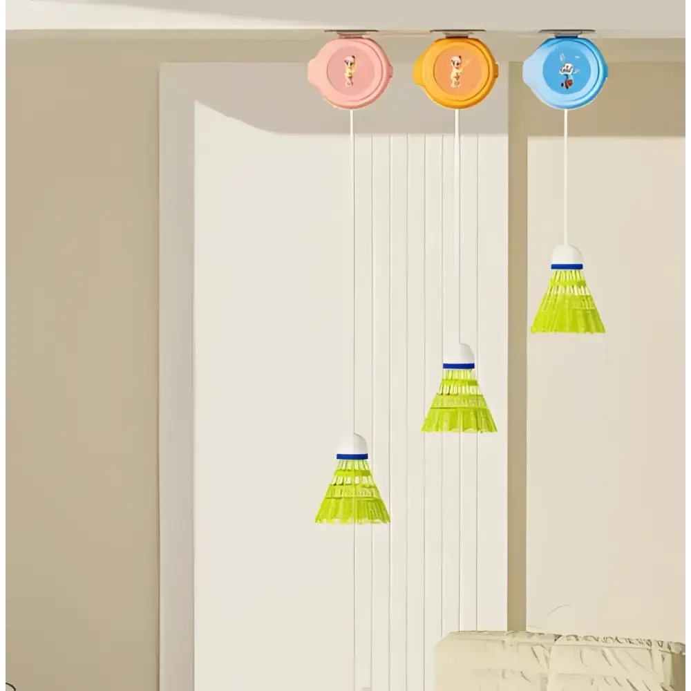 Three colorful indoor badminton trainer shuttlecocks with pastel caps and green feathers hang from orange ceiling holders for hand-eye coordination and racket control