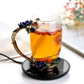 Ornate glass mug cup warmer with gold-toned handle and blue floral design filled with amber tea and goji berries