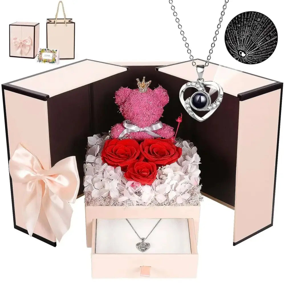 Pink moss bear with heart necklace and gold crown sits on red roses and white hydrangeas in elegant gift box