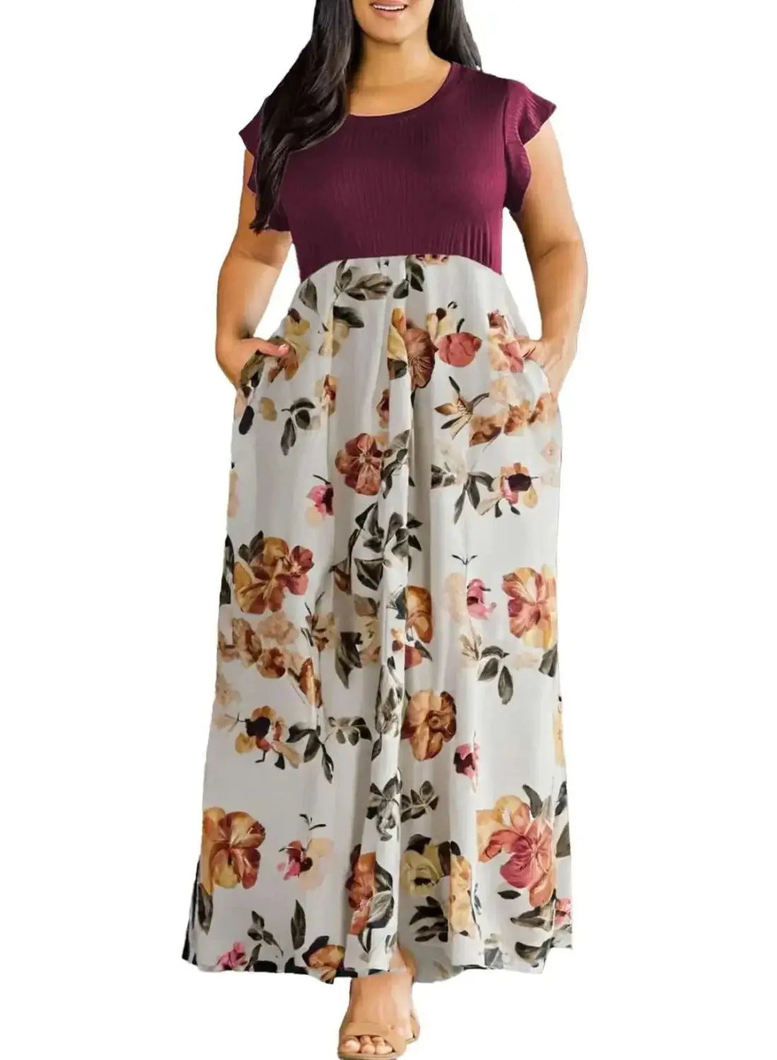 A plus size woman wears a two toned maxi dress with a solid maroon top and a flowing light gray skirt featuring a vibrant floral pattern in orange pink and brown designed for curvy women.