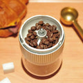 A sleek gold-toned portable electric ceramic coffee grinder with usb charging capability and textured metallic band filled with dark beans
