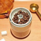 A sleek gold-toned portable electric ceramic coffee grinder with usb charging capability and textured metallic band filled with dark beans