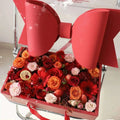 A handheld flower box with a clear window features a vibrant red bow knot and assorted roses gerbera daisies and citrus slices