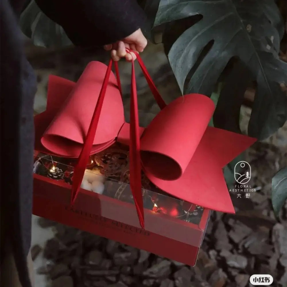 Red bow knot handheld flower box in red paper with rolled ribbon handles and a clear window displaying decorative items