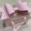 A clear plastic handheld flower box with clear window topped by a large soft pink paper red bow knot