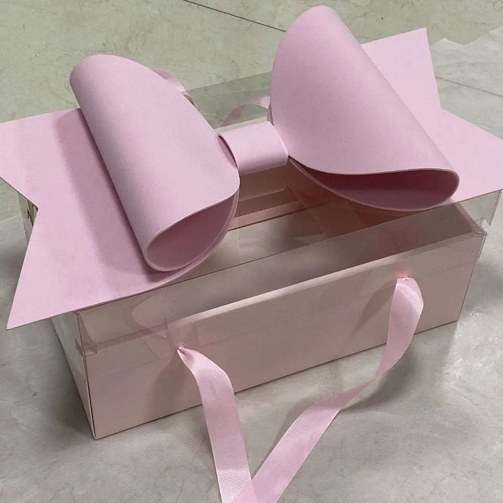 A clear plastic handheld flower box with clear window topped by a large soft pink paper red bow knot