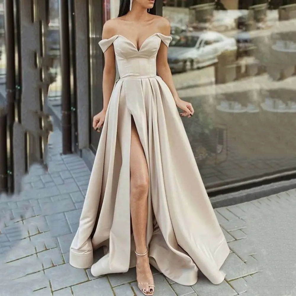 A flowing off-the-shoulder satin gown in soft beige with a high slit and structured bodice, styled as a swing split sleeveless gown