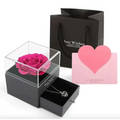 A vibrant pink preserved rose displayed in a clear acrylic box with a black base in a royal blue preserved rose jewelry gift box