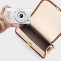 Silver portable ccd digital camera with textured grip and circular lens held above an open beige leather satchel