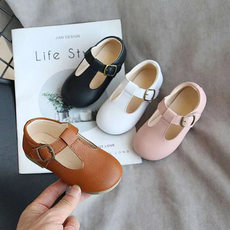 A hand holds a tan leather versatile single shoe toddler mary jane with a gold-toned buckle and rounded toe