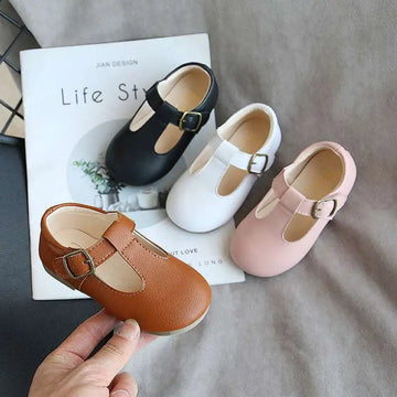 A hand holds a tan leather versatile single shoe toddler mary jane with a gold-toned buckle and rounded toe