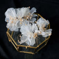 Delicate white organza tulle flower headband and earring set adorned with gold beads displayed in hexagonal glass box