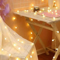 A rustic white wooden folding table with slatted top and x-shaped legs decorated with warm yellow sunshine pearl lights